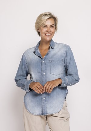 Smiling person with short blonde hair wearing a light blue denim button-up shirt and beige pants, standing against a plain light background.