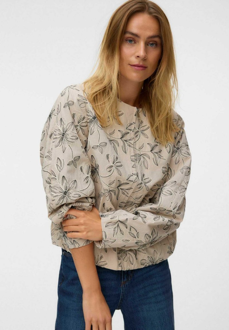 Femme blonde portant un chemisier beige à fleurs à manches longues bouffantes et un jean bleu, posant avec un bras croisé sur son corps.