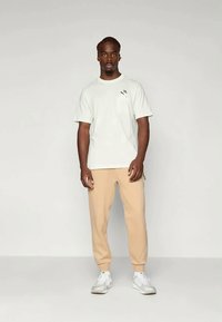 T-shirt de manga curta cinza claro com um pequeno bolso no peito, combinado com calças de moletão bege ajustadas e ténis atléticos brancos.