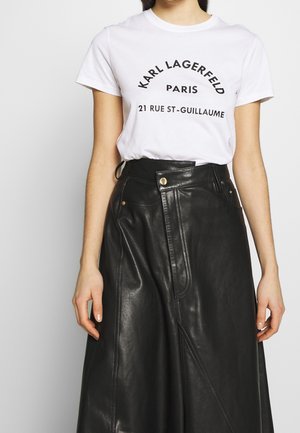 Valkoinen t-paita, jossa musta teksti, sisältäen brändin ja sijainnin. Yhdistetty pitkään mustaan nahkahameeseen, joka on tyylikäs ja rakenne suunniteltu.