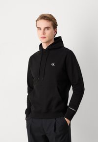 Hoodie noir en coton doux avec une poche avant, un cordon de serrage réglable et un logo blanc Calvin Klein sur la poitrine et la manche.