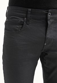 Jeans de mezclilla negra con corte slim, que cuentan con cinco bolsillos, herrajes de metal y una textura suave. Costuras distintivas y acentos de botón.