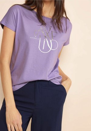 Žena v levandulovém tričku se stříbrným nápisem "NATURAL & LIVE" a tmavě modrých kalhotách, s jednou rukou v kapse, na neutrálním pozadí.