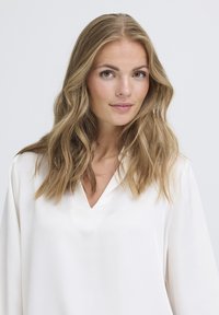 Blusa a maniche lunghe con scollatura a V in morbido tessuto bianco. Texture liscia con design leggero e fluido. Capelli acconciati in onde morbide.