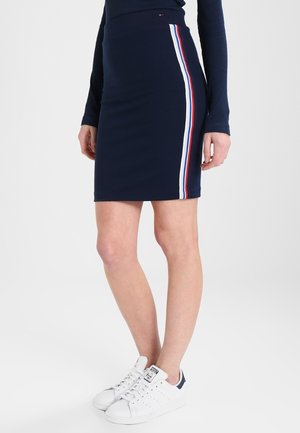 Granatowa dopasowana spódnica z białymi, czerwonymi i niebieskimi paskami po bokach, noszona z granatową bluzką z długim rękawem i białymi trampkami.