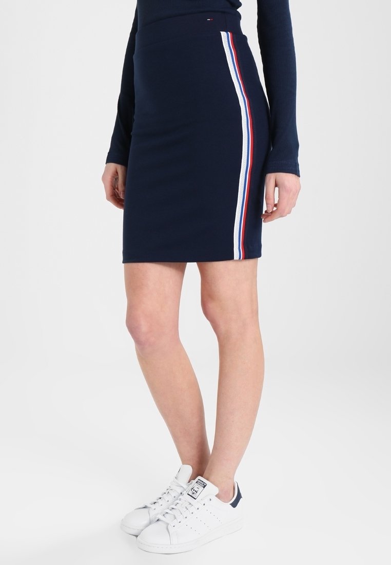 Jupe ajustée bleu marine avec des bandes latérales blanches, rouges et bleues, portée avec un haut à manches longues bleu marine et des baskets blanches.
