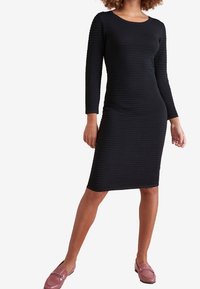 Robe noire côtelée à manches longues, encolure ronde et silhouette ajustée. Associée à des mocassins rose pâle pour un contraste.