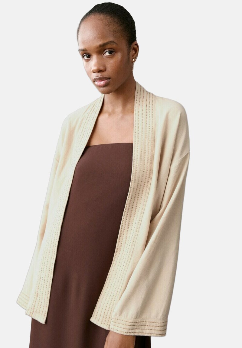 Femme portant une robe marron sans bretelles avec un cardigan ouvert devant, ample, de couleur beige clair, présentant des détails de couture subtils.