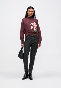 Mujer vestida con un sudadera burdeos con una gran flor rosa, jeans negros, botas negras de tobillo y sosteniendo un pequeño bolso negro.