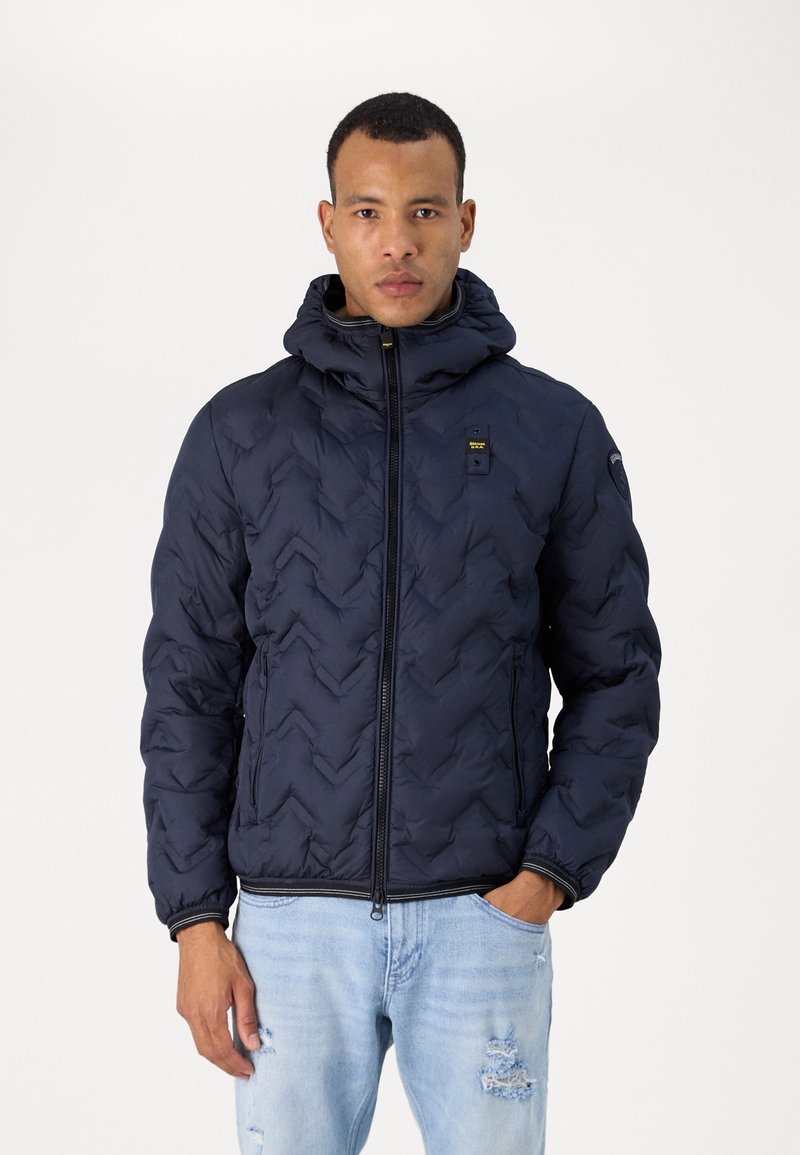Giacca puffer blu navy con motivo a zig-zag, chiusura con zip, cappuccio regolabile e tasche laterali, indossata sopra jeans chiari strappati.