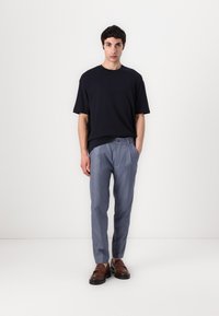 Námořnické bavlněné tričko v kombinaci s světle modrými kalhotami na míru s knoflíkem v pase. Model obouvá hnědé slip-on boty a stojí na neutrálním pozadí.