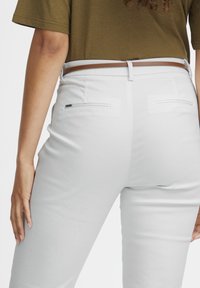 Le dos d'une femme est visible, portant une ceinture marron sur un pantalon blanc, avec sa main gauche posée sur sa cuisse. Le haut est vert olive.