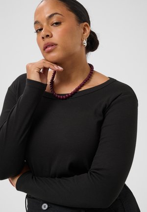 Femme aux cheveux foncés attachés porte un haut noir à manches longues, un collier de perles bordeaux et des boucles d’oreilles en argent en forme d’anneaux, posant avec la main près du visage.