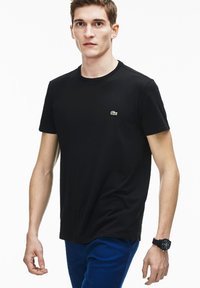 Camiseta de algodón negra con cuello redondo y mangas cortas; presenta un pequeño logotipo bordado en verde en el lado izquierdo del pecho. Modelo que lleva pantalones azules.