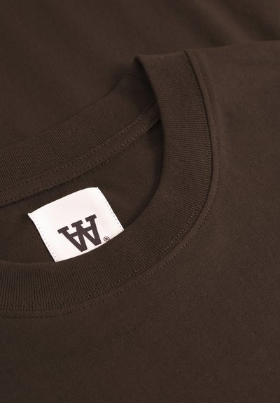 Gros plan sur le col d'un t-shirt noir classique en marron foncé avec une étiquette blanche portant un logo stylisé en noir en double "A" cousu à l'intérieur de l'encolure.