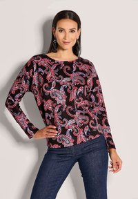 Langärmliges Oberteil aus schwarzem Stoff mit einem lebhaften rot-blauen Paisleymuster. Runder Ausschnitt und lockere Passform. Kombiniert mit dunklem Denim.