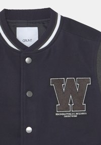 Tmavě modrý univerzitní kabát s bílými a šedými akcenty; má žebrovaný límec, patentní knoflíky a vyšívané logo "W" s nápisem "WASHINGTON D.C. WIZARDS".