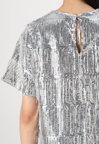 Top in paillettes argentate con maniche corte, caratterizzato da dettagli frangiati, un collo alto e una schiena con apertura a chiave. Texture liscia con riflessi.