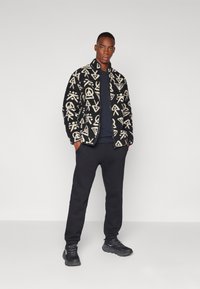Jachetă din fleece neagră și crem cu modele geometrice, cu închidere cu fermoar; combinată cu un hanorac negru și pantaloni jogging, pantofi sport negri.
