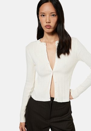 Vrouw met lang donker haar, gekleed in een off-white geribd vest met knopen en zwarte broek, staand met één hand op haar heup.