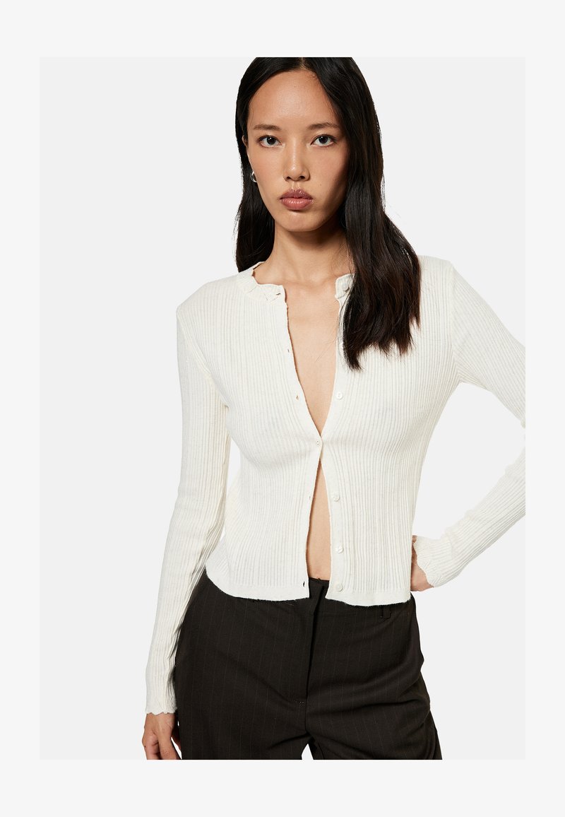 Woman with long dark hair wearing an off-white ribbed buttoned cardigan and black pants, standing with one hand on her hip.