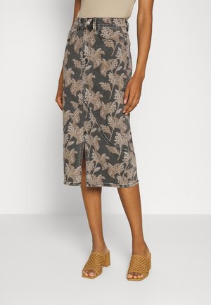 Kvinde iført en højtaljet knælængde nederdel med beige blomsterprint og frontslids, parret med brune vævede højhælede sandaler.