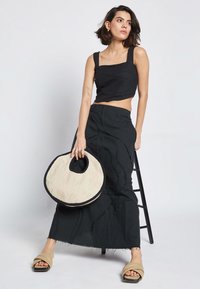 Črn skrajšan top in dolga krila z teksturiranim vzorcem, v kombinaciji z okroglo pleteno torbo in bež sandalami s širokimi trakovi.