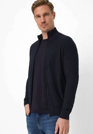 Blond man wearing a dark navy zip-up cardigan over a black shirt and blue jeans, standing with one hand in pocket against white background.