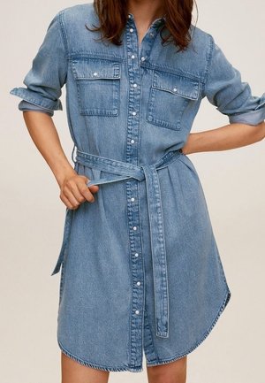 Femme portant une robe-chemise en jean ceinturée avec manches retroussées et poches à rabat à l'avant, les mains ajustant la ceinture.
