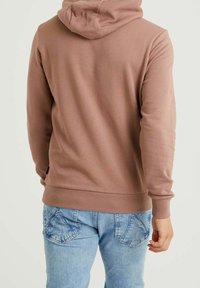 Hnědá mikina s kapucí na stažení, s žebrovanými manžetami a spodním lemem, v kombinaci se světle modrými džíny z denimu s charakteristickým prošíváním na kapsách.