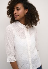 Blusa bianca con colletto alto, maniche lunghe e motivo ad occhiello. Presenta bottoni e un orlo arricciato con un tessuto morbido e testurizzato.