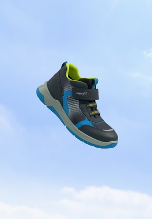 Børnesportssneaker i sort, blå og neongrøn med velcrolukning og elastiske snørebånd mod en klar blå himmel.