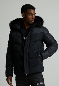 Svart puffer-jakke med fleece-fôret hette, med en kviltet tekstur og glidelås. Utsmykket med geometriske mønstre på skuldrene.