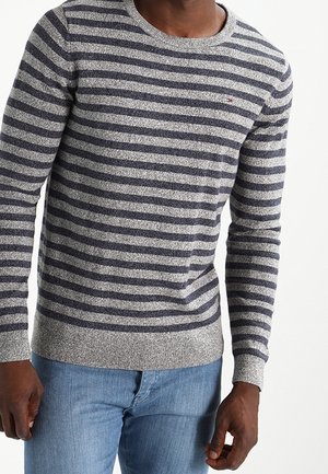 Mann trägt einen grau-schwarz horizontal gestreiften Pullover und blaue Jeans, steht vor einem schlichten weißen Hintergrund.