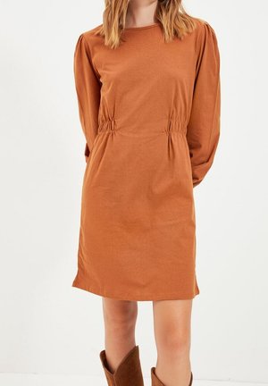 Vrouw draagt een lange mouwen, roestkleurige jurk met ingesnoerde taille en bruine halfhoge laarzen, staand tegen een effen witte achtergrond.