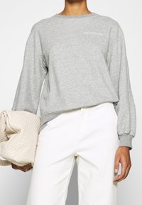 Harmaa collegepaita pyöreällä pääntiellä ja pitkällä hihalla, valmistettu pehmeästä kankaasta. Rinnassa pieni valkoinen logo, yhdistetty vaaleanruskeisiin housuihin.