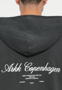 Grauer Kapuzenpulli mit weicher Textur, großer Kapuze und weißem Schriftzug "Arkk Copenhagen" auf dem Rücken mit zusätzlichem Text darunter.