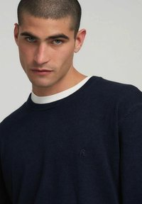 Maglione blu navy realizzato in tessuto morbido, caratterizzato da una scollatura rotonda con uno strato interno bianco e un piccolo logo nell'angolo inferiore.