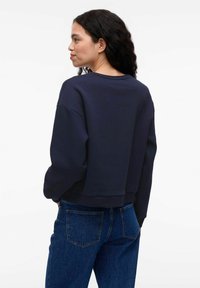 Tamsiai mėlynas trumpas džemperis su ilgomis rankovėmis ir apvaliu iškirpimu. Minkšta tekstūra, su raukiniais ant rankovių ir apačios, derinamas su mėlynos džinsinės medžiagos kelnėmis.