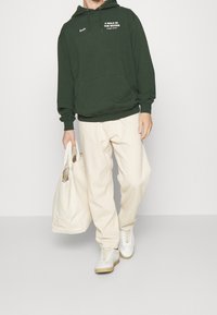 Felpa verde scuro con tasca frontale, testo stampato bianco. Pantaloni beige larghi, che portano una borsa in tessuto leggera. Sneakers bianche con suole beige.
