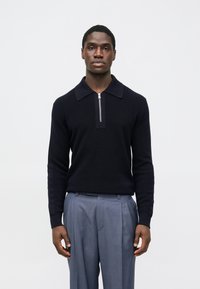 Námornícky modrý pletený sveter s golierovým výstrihom a polovičným zipsom, kombinovaný s ľahkými sivými plisovanými nohavicami. Hladká textúra a priliehavý dizajn.