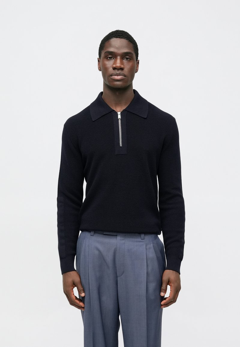 Námornícky modrý pletený sveter s golierovým výstrihom a polovičným zipsom, kombinovaný s ľahkými sivými plisovanými nohavicami. Hladká textúra a priliehavý dizajn.