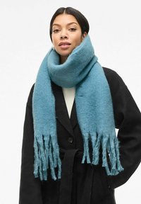 Turkusowy wełniany szalik z detalami z frędzlami, zestawiony z czarnym płaszczem. Miękka faktura, wygodnie układa się na szyi.
