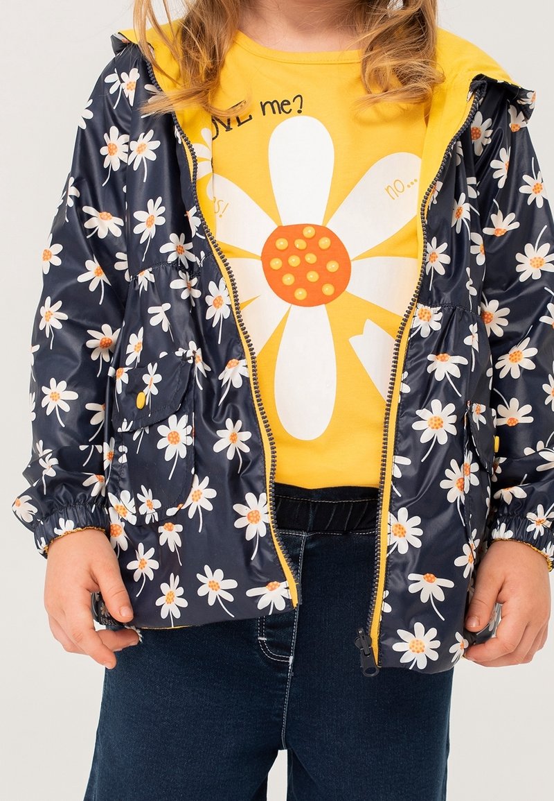 Enfant portant une veste bleu marine avec des marguerites blanches sur un t-shirt jaune avec une grande marguerite blanche et un pantalon bleu.