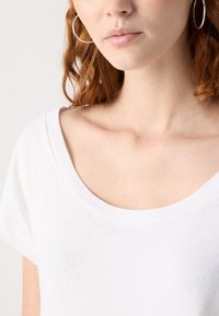 Jeune personne aux cheveux roux portant de grandes boucles d’oreilles anneaux et un t-shirt blanc à col rond sur un fond uni.