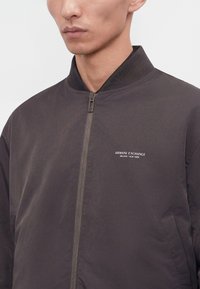 Homme portant une veste bomber zippée gris foncé avec le logo « Armani Exchange Milano / New York » sur la poitrine gauche.