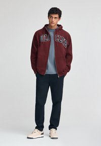 Vinröd zip-up hoodie med texten "POLO CLUB", grå skjorta under, svarta byxor och ljusa sneakers. Hoodien har dragsko och kengurufickor.
