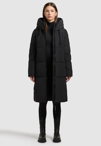 Sort polstret lang frakke med hætte, der har frontlynlås og lodrette syninger. Glat tekstur og struktureret design.