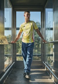 Geel en witte t-shirt met korte mouwen met roze accenten, blauwe jeans en zwarte schoenen; staand in een glazen lift met metalen vloer.