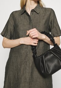 Donna che indossa un abito a maniche corte di colore verde oliva con bottoni, mentre tiene con entrambe le mani una piccola borsa in pelle arricciata di colore nero.